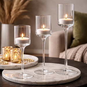 Three clear glass candle holders with long stems are arranged on a round white marble tray on a dark table. Each holder contains a white tea light candle, with two candles lit and casting a warm glow.