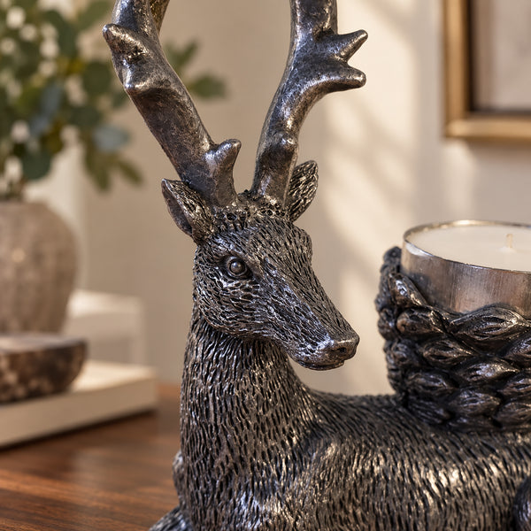 A metallic deer figurine with large antlers, shown in a home setting on a wooden table next to a braided candle holder with a silver candle.