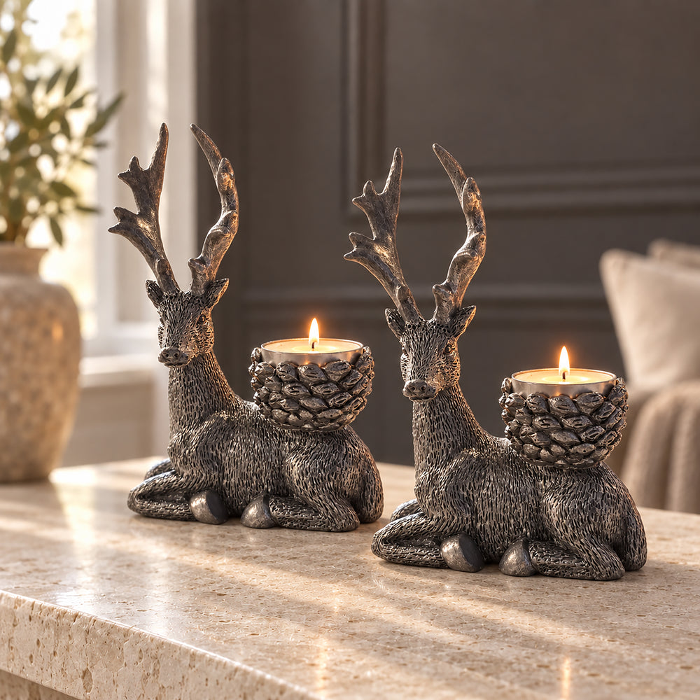 Two metallic deer candle holders with antlers and pinecone-shaped cups holding lit tealight candles, displayed on a stone tabletop in a softly lit interior setting.