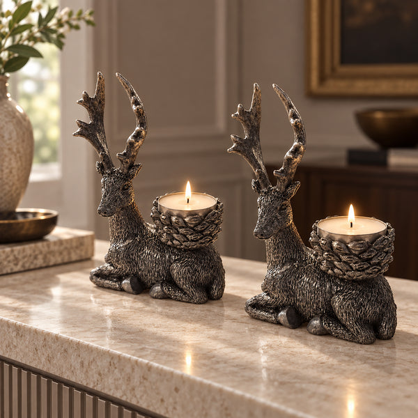 Two deer-shaped candle holders with antlers and textured bodies sit on a marble countertop in a warmly lit interior setting. Each deer holds a lit tealight candle, with a vase and framed decor softly blurred in the background.