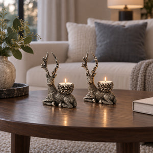 Two metallic deer candle holders with textured silver and gold finishes, each holding a lit tea light, displayed on a dark wooden coffee table in a cozy living room setting.