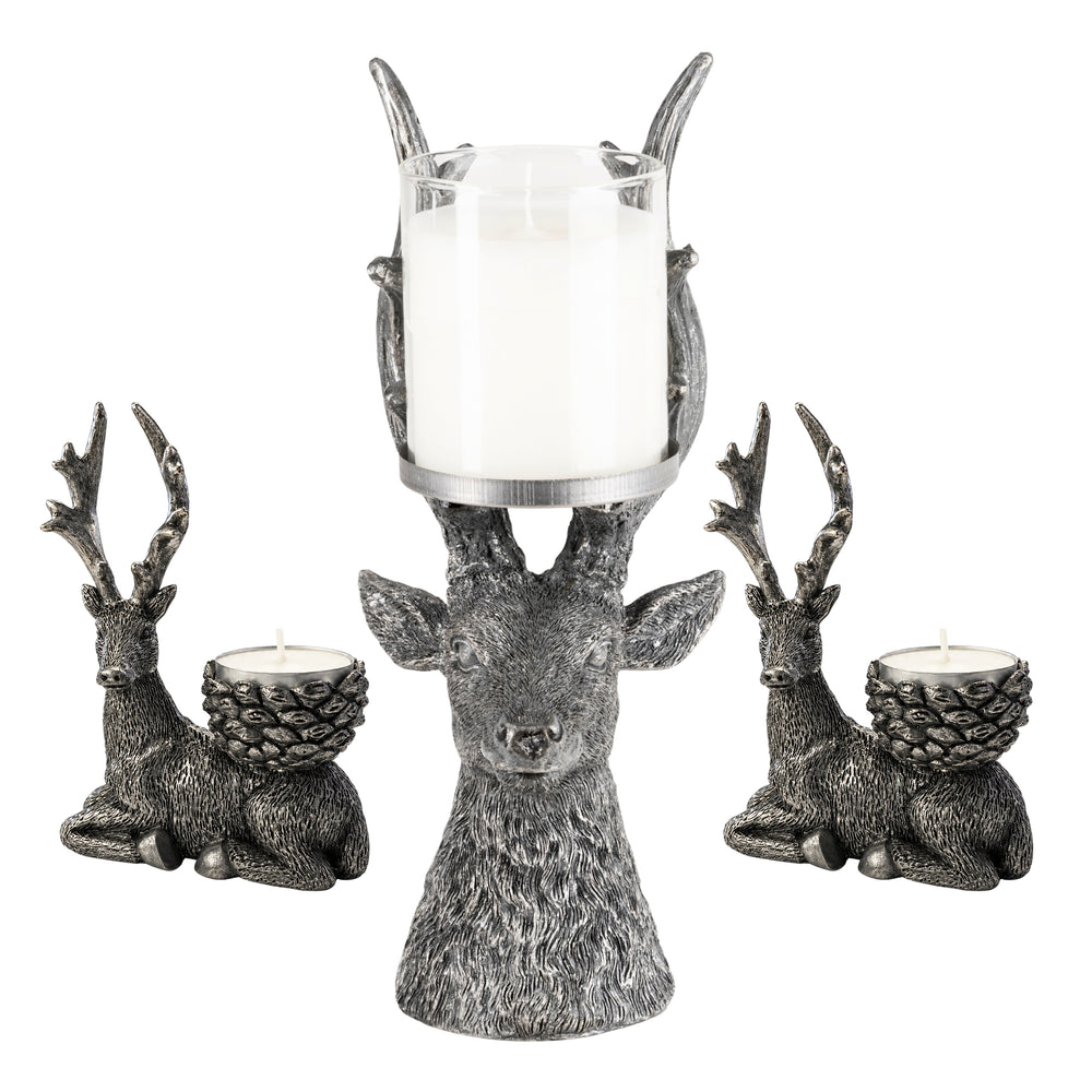 Three deer-shaped candle holders in a distressed metallic finish are arranged on a light stone surface. The center deer holds a tall glass candle, while the two side deer each hold a small tea light candle.