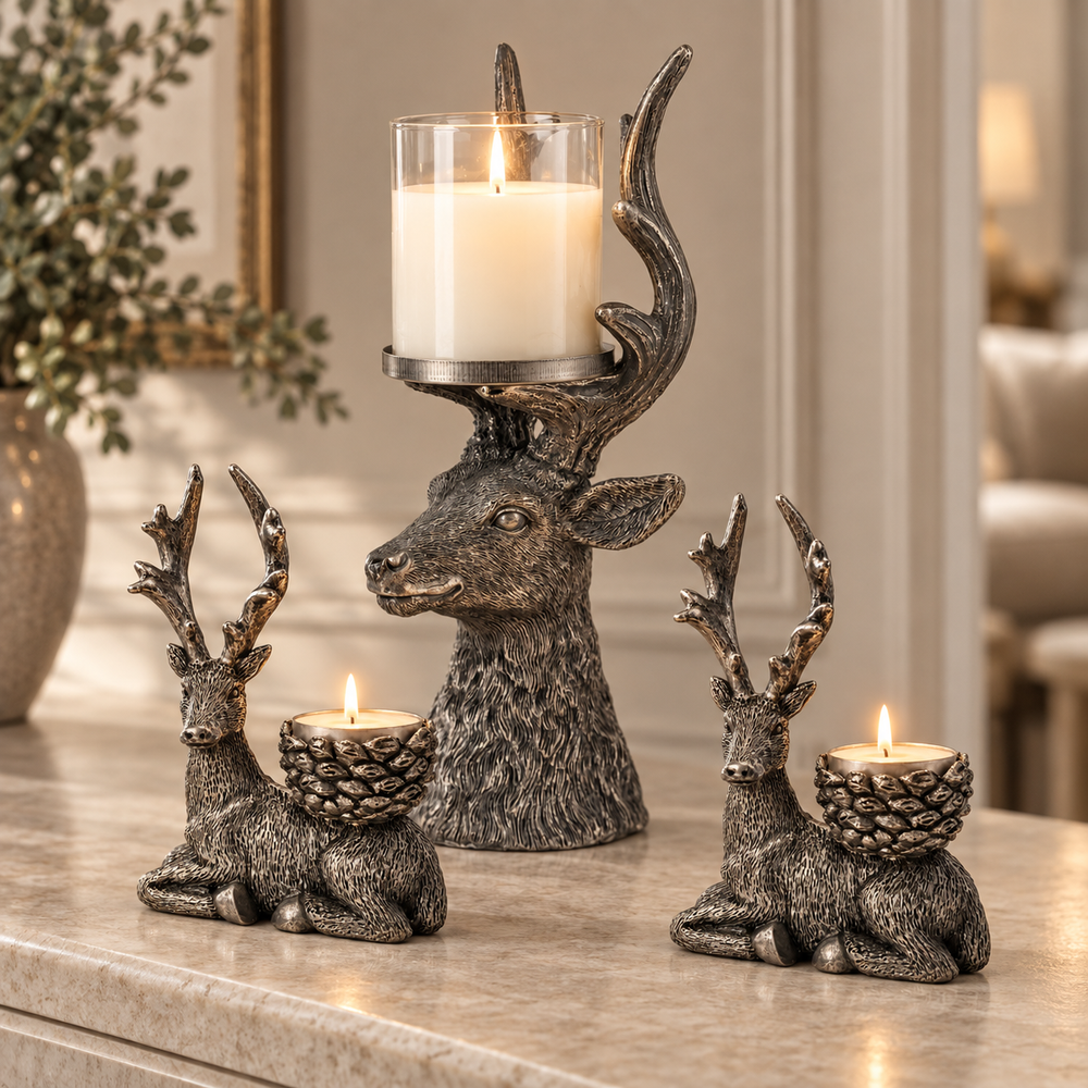 Three deer-shaped candle holders in a distressed metallic finish are arranged on a light stone surface. The center deer holds a tall glass candle, while the two side deer each hold a small tea light candle.