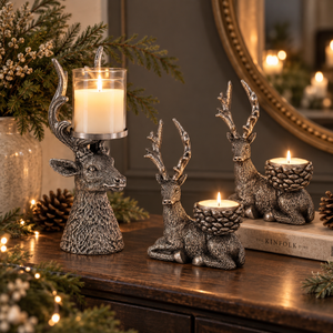 Three metallic deer-shaped candle holders with antlers are arranged on a wooden table in a decorated indoor setting. One stands upright holding a tall glass candle, one reclines holding a tea light, and one sits on a book holding a candle.