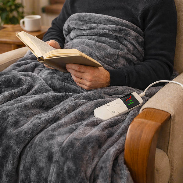 A person sitting in a beige armchair, wrapped in a plush gray blanket while reading a book. A white wired digital temperature controller rests on the blanket next to them, with a mug on a table in the background.