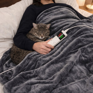 A gray plush blanket covers a person in bed, with a brown tabby cat resting on their chest. The person holds a white digital temperature controller connected to the blanket, which is shown regulating the blanket's temperature in a cozy bedroom setting.