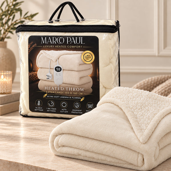 A folded cream-colored heated throw blanket is shown in the foreground, with its branded packaging bag standing behind it on a light stone surface. The package reads 'MARCO PAUL' and 'HEATED THROW', and the scene is styled in a warm indoor setting with soft lighting and home decor in the background.