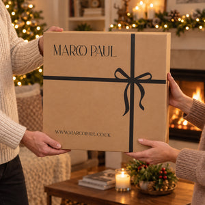 A large brown cardboard gift box with 'MARCO PAUL' printed on the front, held by two people in a cozy holiday living room with a fireplace and Christmas lights in the background.
