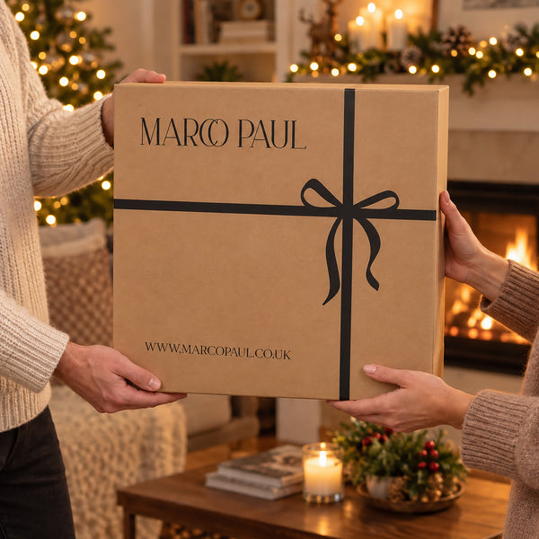 A large brown cardboard gift box with 'MARCO PAUL' printed on the front, held by two people in a cozy holiday living room with a fireplace and Christmas lights in the background.