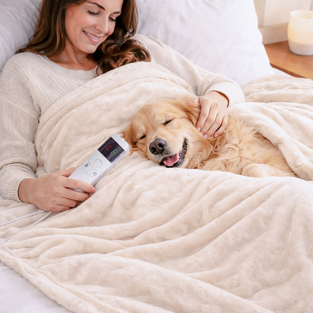 A woman and her golden retriever dog relax under a cream-colored heated blanket on a bed, with the woman holding a white remote control.
