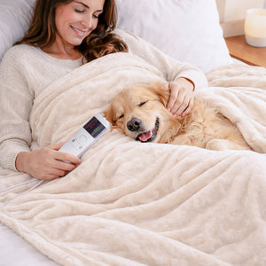 A woman and her golden retriever dog relax under a cream-colored heated blanket on a bed, with the woman holding a white remote control.