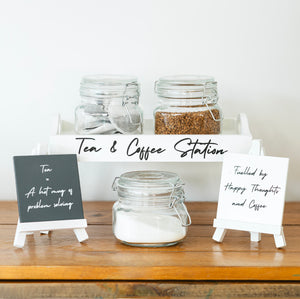 Tea and coffee station with jars and small signs on a wooden surface.