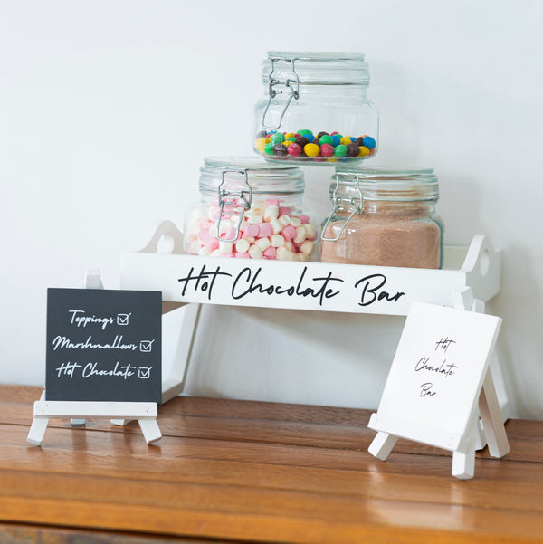 Hot chocolate bar setup with jars of toppings on a wooden surface.