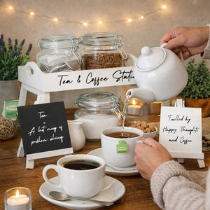 A cozy tabletop scene showing a white teapot pouring hot water into a cup of tea. A hand holds the teapot, and another hand holds a cup with a visible Green Tea tea bag tag.