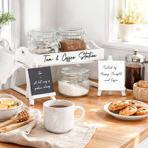 A styled tea and coffee station setup on a wooden table, featuring glass jars with contents, a white mug of coffee, cookies on a plate, and small signboards reading 'Tea & Coffee Station' and 'Fueled by Happy Thoughts and Coffee.'.