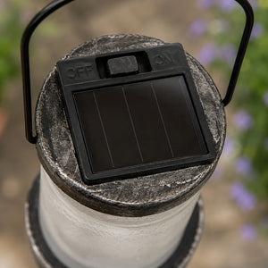 Close-up of a solar-powered outdoor lantern showing its black solar panel cover with ON/OFF switches, a textured gray top, and a white cylindrical body in an outdoor garden setting.