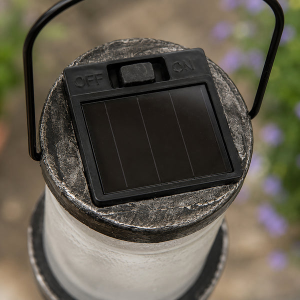 Close-up of a solar-powered outdoor lantern showing its black solar panel cover with ON/OFF switches, a textured gray top, and a white cylindrical body in an outdoor garden setting.