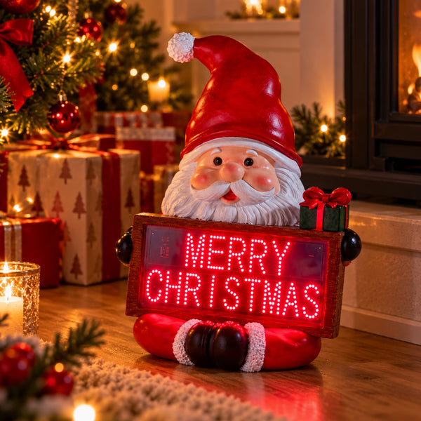 A festive Santa Claus figurine holding a glowing red LED sign that reads 'MERRY CHRISTMAS,' placed on a wooden floor in a decorated holiday home with wrapped gifts, a Christmas tree, and warm lights in the background.