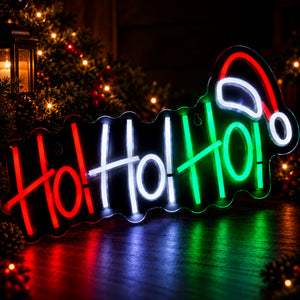 A decorative neon-style sign spelling "Ho Ho Ho" in red, white, and green, with a red Santa hat at the top, photographed in a festive holiday setting with lights and pine branches in the background.
