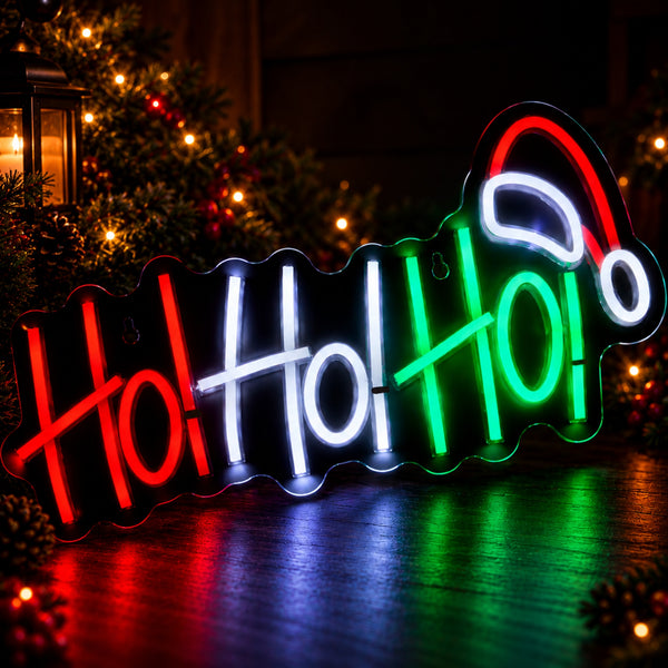 A decorative neon-style sign spelling "Ho Ho Ho" in red, white, and green, with a red Santa hat at the top, photographed in a festive holiday setting with lights and pine branches in the background.