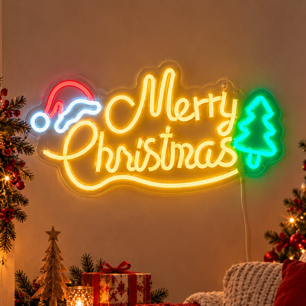 A glowing Christmas neon sign on a warm beige wall, featuring the words “Merry Christmas” in yellow script with a red Santa hat on the left and a green Christmas tree on the right. The sign is styled in a cozy holiday setting with Christmas trees, wrapped gifts, and a knitted blanket in the foreground.