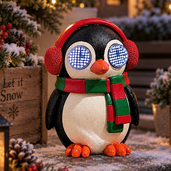 A penguin-shaped decorative figure wearing red earmuffs and a red-and-green striped scarf, standing outdoors in a snowy setting with warm holiday lights and evergreen branches in the background.