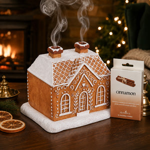 A gingerbread house-shaped smoking confectionery toy or decor piece on a wooden table beside its product box labeled 'cinnamon' by Cleer & Co. The scene is styled with a warm fireplace, blurred Christmas lights, dried orange slices, and greenery in the background.