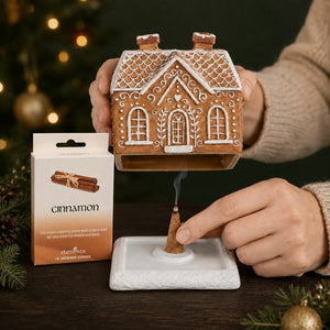 A ceramic gingerbread house-shaped incense holder is shown with a burning incense stick placed in its base. A person's hands are visible holding the house and lighting the incense.