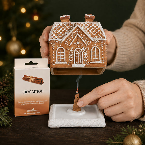 A ceramic gingerbread house-shaped incense holder is shown with a burning incense stick placed in its base. A person's hands are visible holding the house and lighting the incense.
