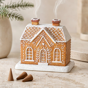 A gingerbread house-shaped incense holder with white icing details, set on a light stone surface with three small incense cones nearby and a softly blurred holiday background.