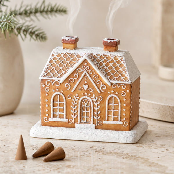 A gingerbread house-shaped incense holder with white icing details, set on a light stone surface with three small incense cones nearby and a softly blurred holiday background.