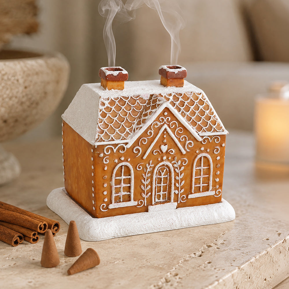 A gingerbread house-shaped candle holder or decorative home candle with white icing-style trim, a snow-dusted roof, two smoking chimneys, and a white base, styled on a tabletop with cinnamon sticks and a warm blurred background.