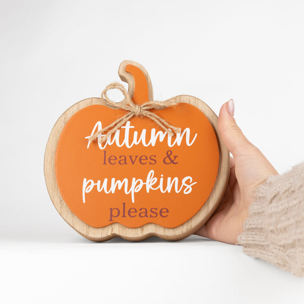 A hand holds a round wooden pumpkin-shaped sign with a bright orange face, natural wood border, jute hanging loop, and white text reading 'Autumn leaves & pumpkins please' on a clean white background.