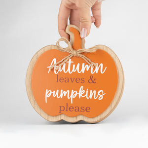 A hand holds a scalloped wooden pumpkin sign with an orange face, natural wood border, jute twine bow, and the phrase 'Autumn leaves & pumpkins please' in white and muted mauve lettering, on a light gray background.