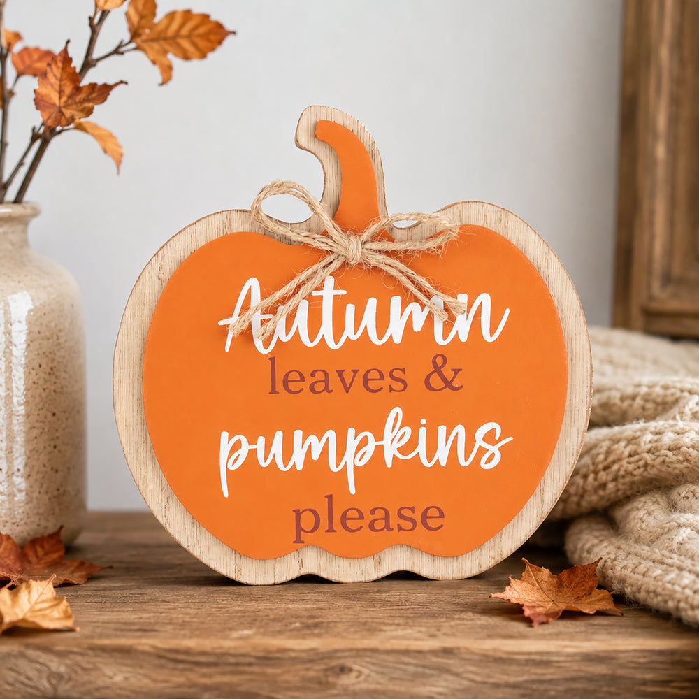 A pumpkin-shaped wooden sign with a bright orange face, natural wood border, and jute bow, reading 'Autumn leaves & pumpkins please' in white and muted red lettering. It sits on a rustic wooden surface with fall leaves, a ceramic vase, and a knitted blanket in the background.