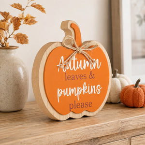 A scalloped wooden pumpkin sign with an orange face and white text reading 'Autumn leaves & pumpkins please,' tied with a jute bow, displayed on a wooden surface with fall decor in the background.