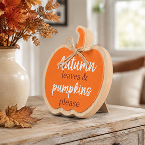 A decorative pumpkin-shaped wooden sign with a bright orange face, natural wood edges, and a small jute bow, standing on a rustic table. The sign reads “Autumn leaves & pumpkins please” in white and dark lettering.