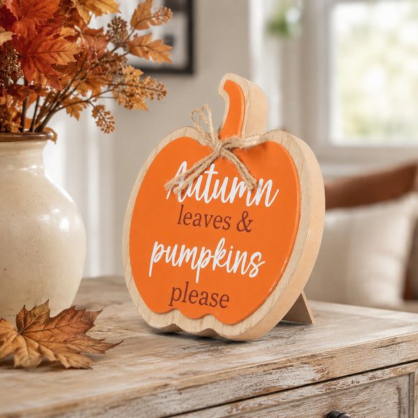 A decorative pumpkin-shaped wooden sign with a bright orange face, natural wood edges, and a small jute bow, standing on a rustic table. The sign reads “Autumn leaves & pumpkins please” in white and dark lettering.