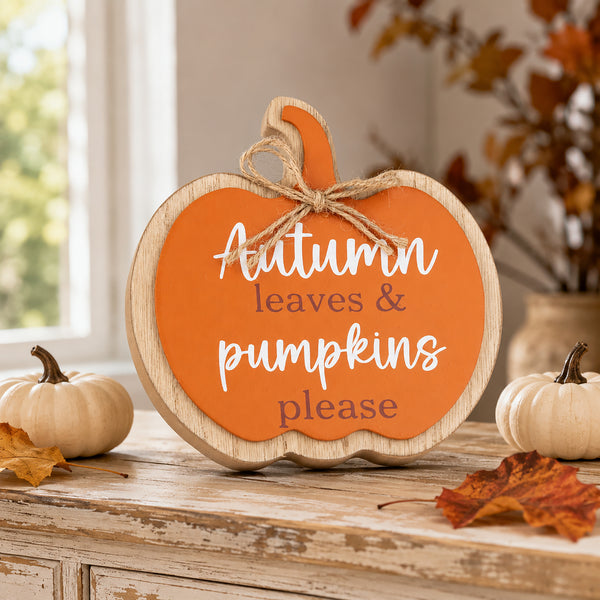 A pumpkin-shaped wooden sign with an orange face, natural wood border, and jute bow, reading “Autumn leaves & pumpkins please,” displayed on a rustic table with fall decor.