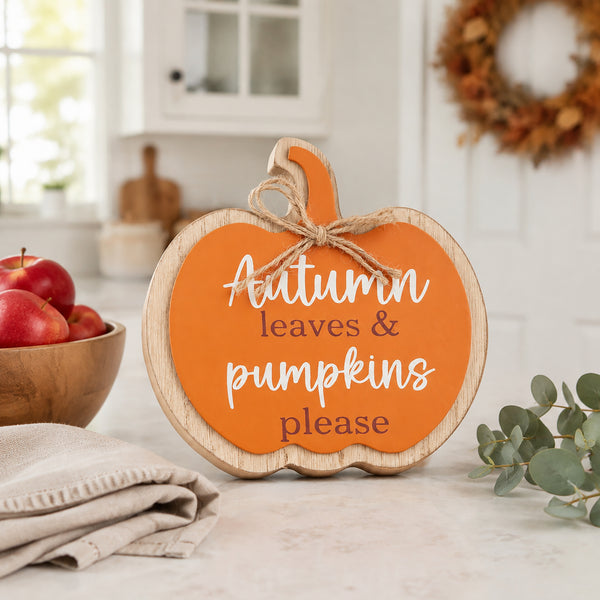 A pumpkin-shaped wooden sign with a bright orange face and natural wood edges, tied with a jute bow. The sign reads 'Autumn leaves & pumpkins please' in white and dark brown lettering.