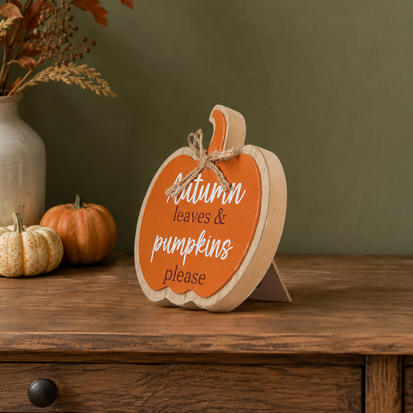 A wooden pumpkin-shaped decorative sign with an orange face and natural wood edges, tied with twine and reading 'Autumn leaves & pumpkins please,' displayed on a wooden table with small pumpkins and dried flowers in the background.