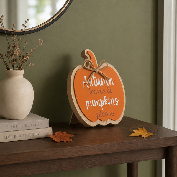 A decorative wooden pumpkin sign with the phrase 'Autumn leaves & pumpkins please' in white script, displayed on a dark wood console table in a styled home setting with a vase, books, and autumn leaves.