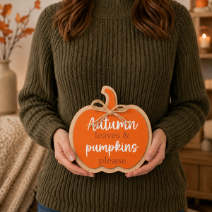 A person wearing an olive green ribbed sweater holds a decorative autumn pumpkin sign that reads 'Autumn leaves & pumpkins please' in white script and serif text on an orange background.
