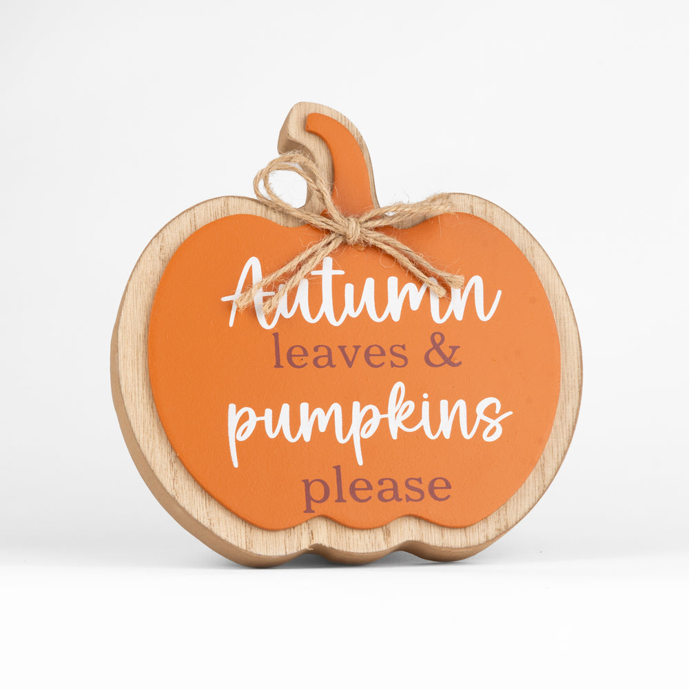 A pumpkin-shaped wooden sign with a bright orange face, natural wood border, and jute bow, reading 'Autumn leaves & pumpkins please' in white and muted red lettering. It sits on a rustic wooden surface with fall leaves, a ceramic vase, and a knitted blanket in the background.