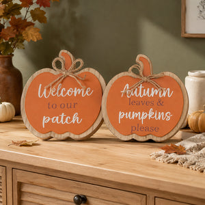 Two decorative pumpkin-shaped wooden signs with orange fronts and natural wood edges, placed on a wooden cabinet in a fall-themed room. One sign reads 'Welcome to our patch' and the other reads 'Autumn leaves & pumpkins please'.
