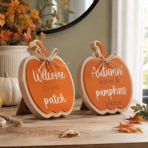 Two pumpkin-shaped wooden signs with orange fronts and white script text, displayed on a wooden table in a fall-themed setting with autumn leaves and a small pumpkin decor piece.