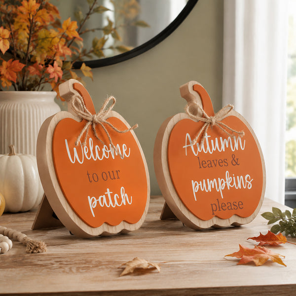 Two pumpkin-shaped wooden signs with orange fronts and white script text, displayed on a wooden table in a fall-themed setting with autumn leaves and a small pumpkin decor piece.