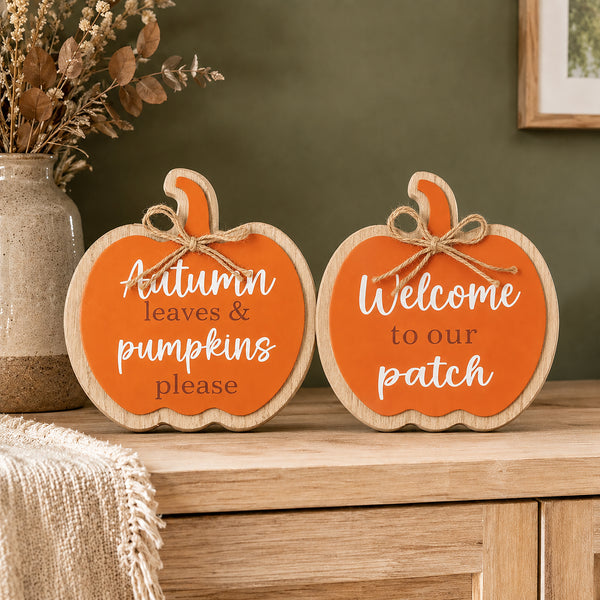 Two pumpkin-shaped wooden signs with orange fronts and natural wood edges sit on a wooden cabinet. One sign reads "Autumn leaves & pumpkins please" and the other reads "Welcome to our patch" in white script.