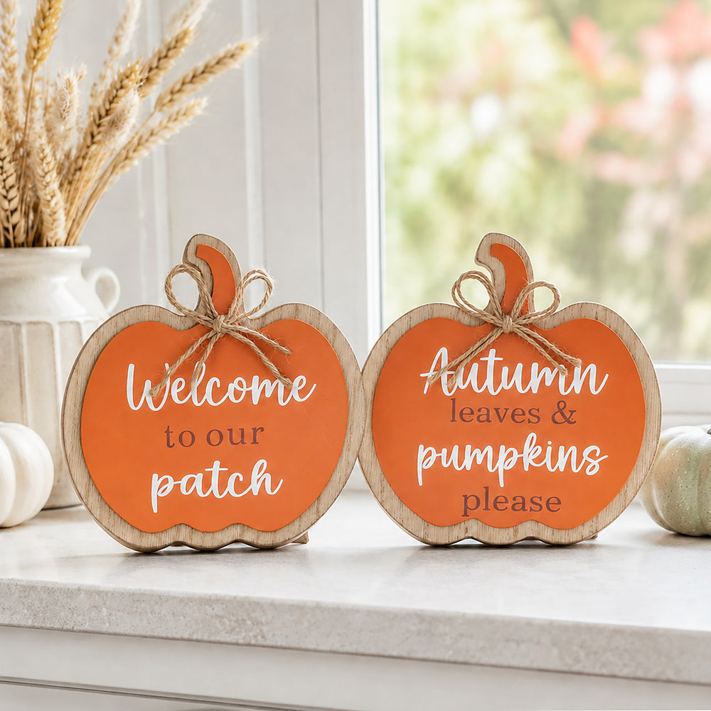 Two pumpkin-shaped wooden signs with orange centers and white lettering, displayed on a windowsill with fall decor. One sign reads "Welcome to our patch" and the other reads "Autumn leaves & pumpkins please.