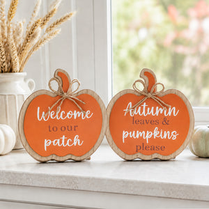 Two pumpkin-shaped wooden signs with orange centers and white lettering, displayed on a windowsill with fall decor. One sign reads "Welcome to our patch" and the other reads "Autumn leaves & pumpkins please.